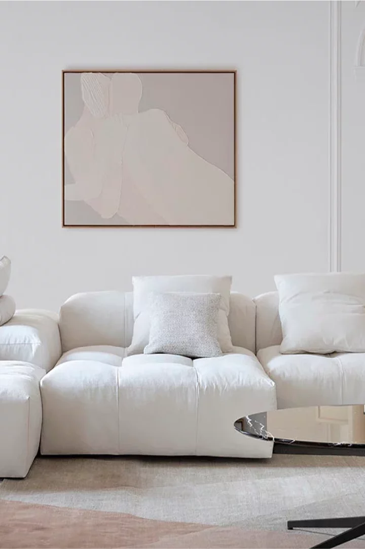 Immagine di un salotto di Saba, cliente di Extera che ha implementato un Dam. L'immagine mostra un divano bianco molto ampio con cuscini, sotto un tappeto dai colori tenui sul beige e il rosa e un quadro sulla parete dietro il divano con un disegno stratto.